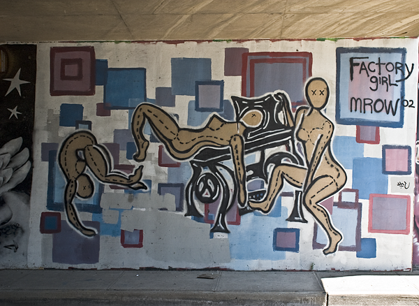 Colorful hand-painted mural title Factory Girl featuring stylized human figures and geometric shapes beneath a concrete overpass.